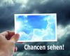 Motiv 2016: Es zeigt einen grauen, wolkigen Himmel. Im Vordergrund hält eine Hand vom linken unteren Bildrand her eine Postkarte in die Bildmitte. Die Karte zeigt einen blauen Himmel mit weißen Wolken. Unter der Postkarte steht das Motto „Chancen sehe