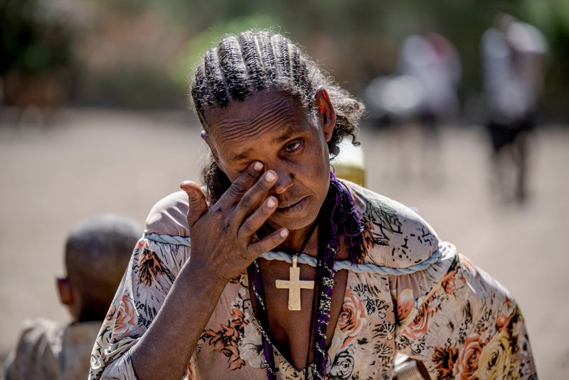 Eine müde aussehende afrikanische Frau mit typisch geflochtenem Haar reibt sich die Augen. Um die Schultern trägt sie ein straffes Seil, an dem sie einen Wasserkanister auf ihrem Rücken trägt. An dem Seil hängt vorne ein großes Kreuzsymbol. 